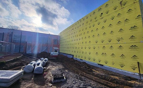 两层实心墙与黄色护套相连，旁边是一栋垂直的塑料建筑. 前景是泥土和建筑设备