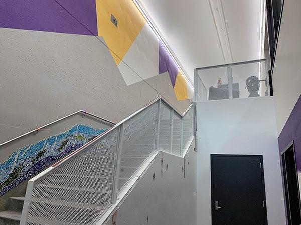 楼梯上有金属网栏杆. 墙的顶部被漆成一片片紫色, gold, 和白色的, 墙上的一幅壁画沿着楼梯向上延伸