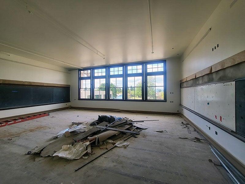 教室左边的墙上有白板, 窗户在远处的墙上, 右边的墙上有一块黑板. 地板上覆盖着施工保护，地板中间有一堆材料