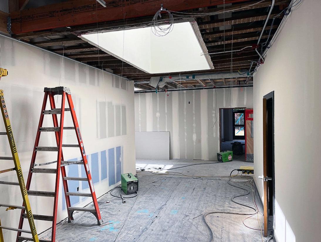 a hallway has wallboard installed and taped, an open ceiling with a skylight, and covered floors. Two ladders are in the lower right and power cords are along the floor.